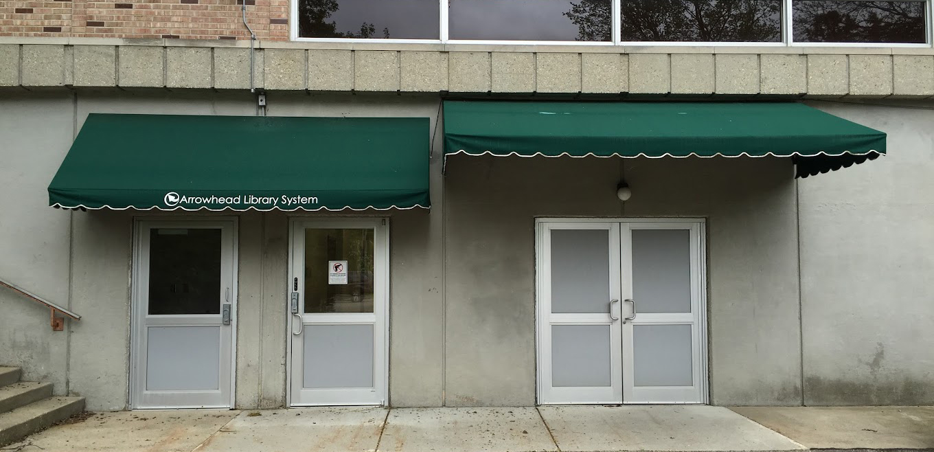 Four metal exterior doors under dark green awnings located in the basement of the Milton Public Library off the back parking lot.
