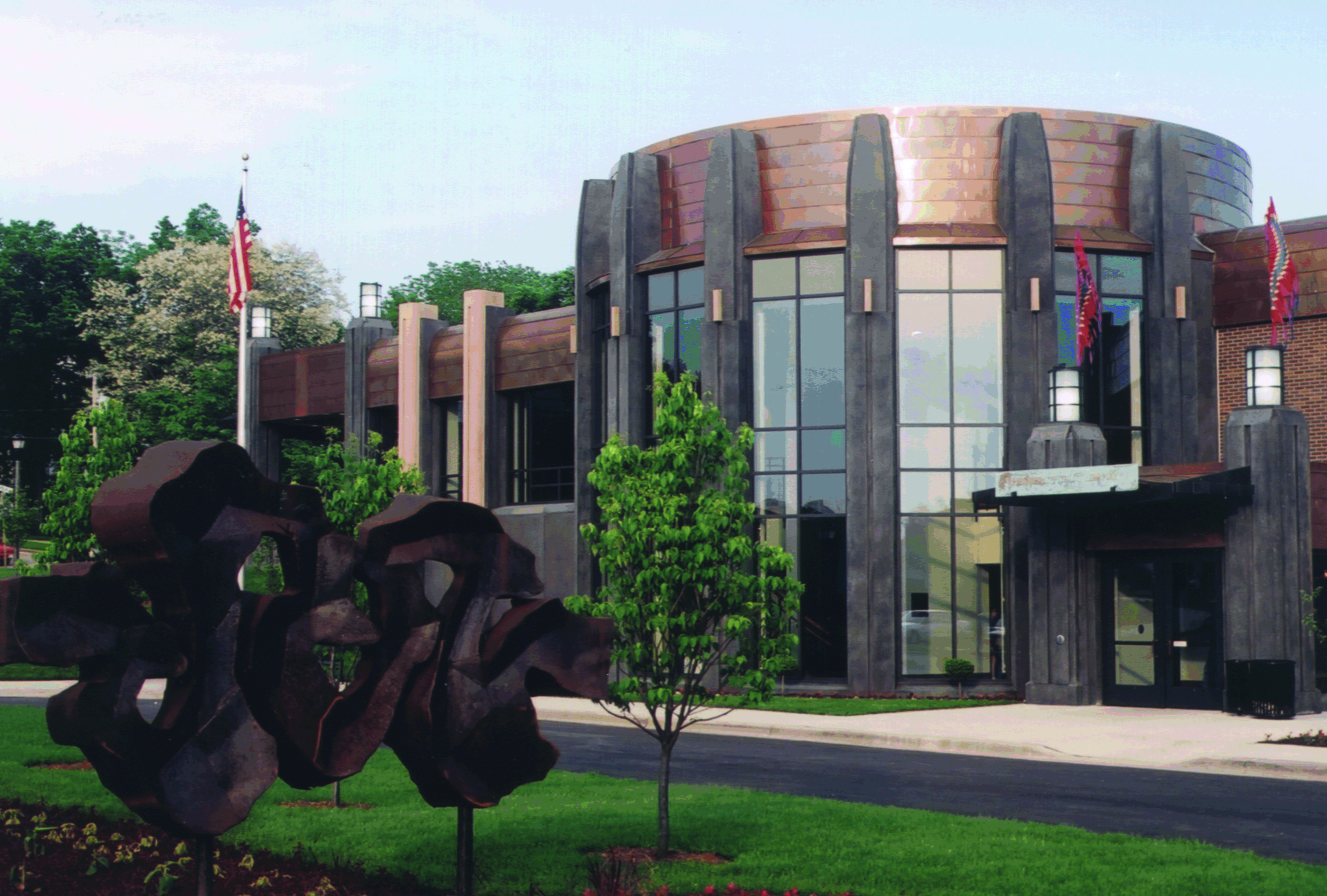 Exterior photo of Hedberg Public Library. The building is brown brick, glass, and copper metal accents. The main portion of the library from this view is a glass and copper rotunda. In the lawn in front of the building is a large abstract sculpture.