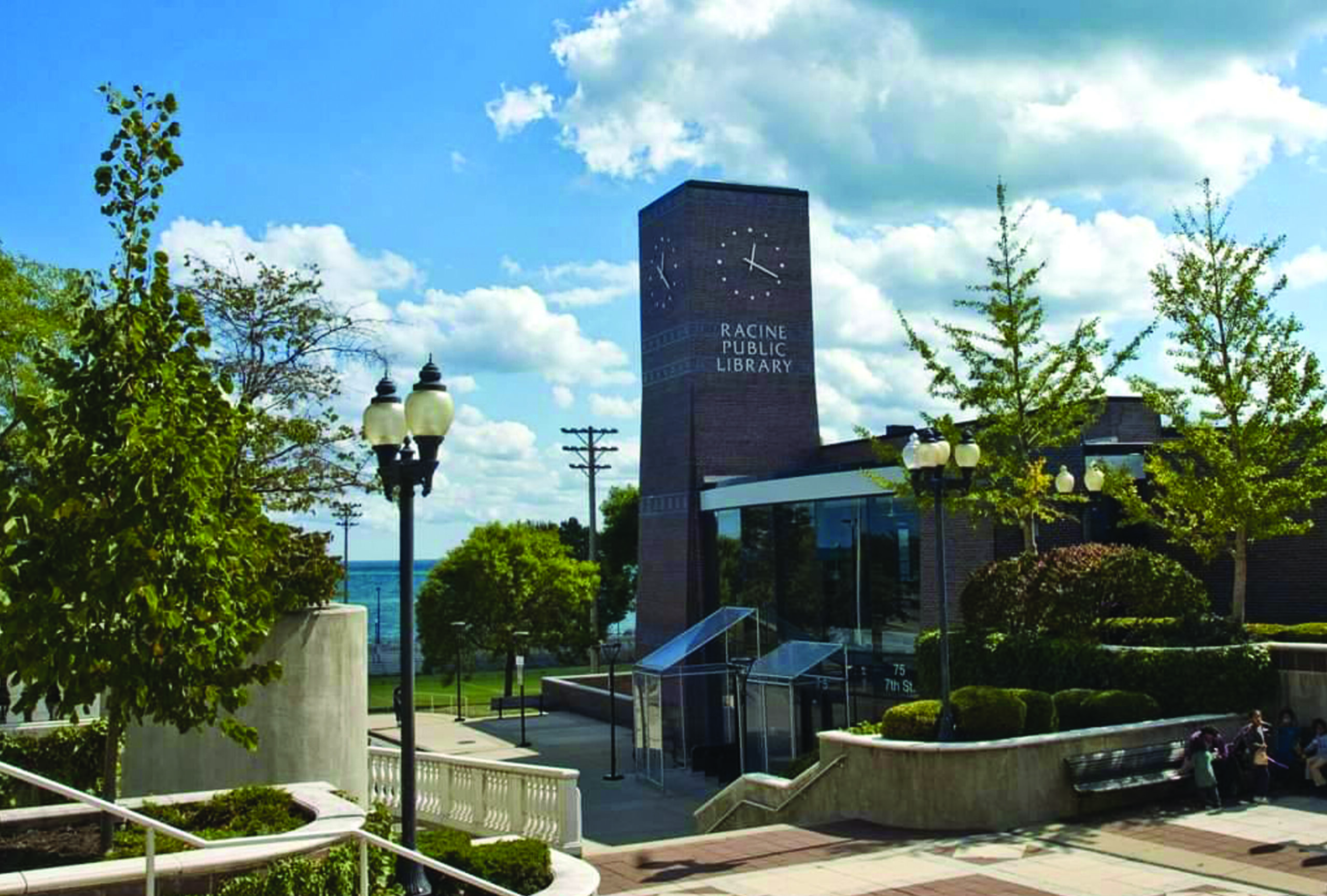 Exterior photo of Racine Public Library. A brick and glass building, featuring a brick clock tower, is nestled in downtown Racine on the shore of beautiful Lake Michigan. The grass and trees are lush and green, and a peek of the lake is visible.