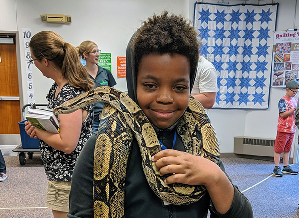 A boy poses in a library program room with a large snake draped around his neck and shoulders.