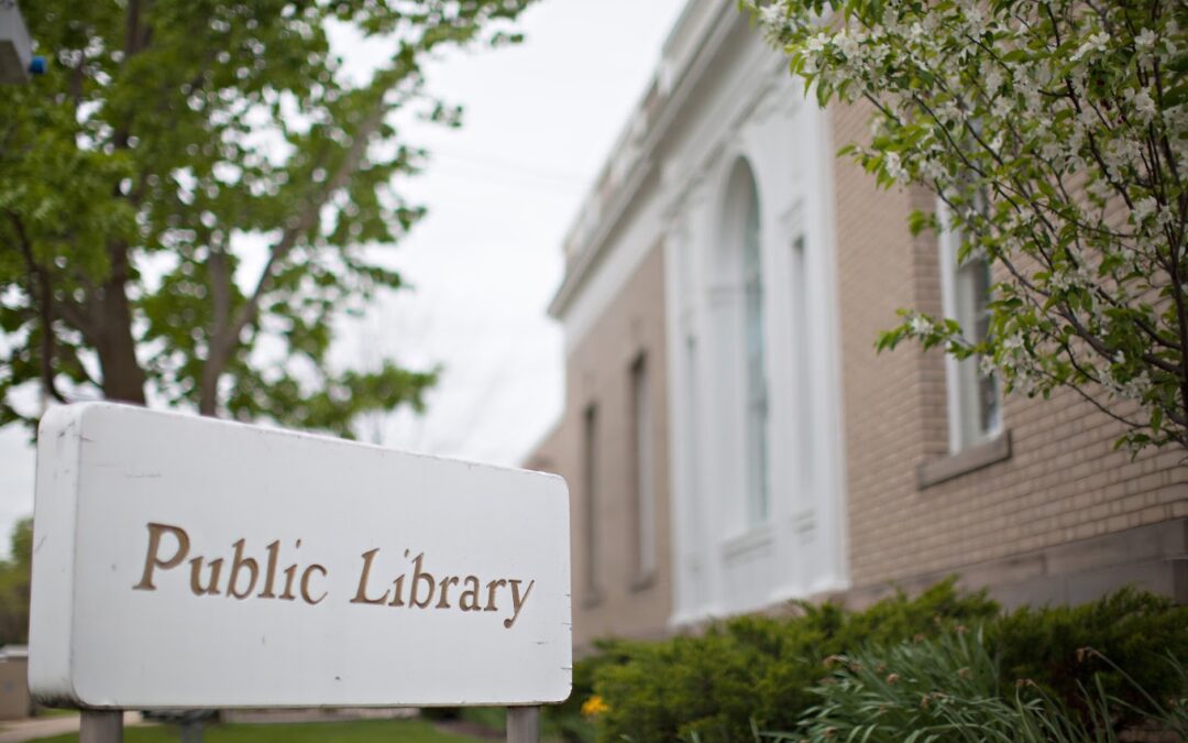The tan brick and white portico of the Burlington Public Library are seen, slightly out of focus in the background. The main focus of the image is on an white outdoor sign reading, "Public Library".