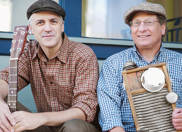 Two men sit outside a storefront. Both wear newsboy caps and fine plaid shirts. One holds a string instrument and one holds a washboard instrument.