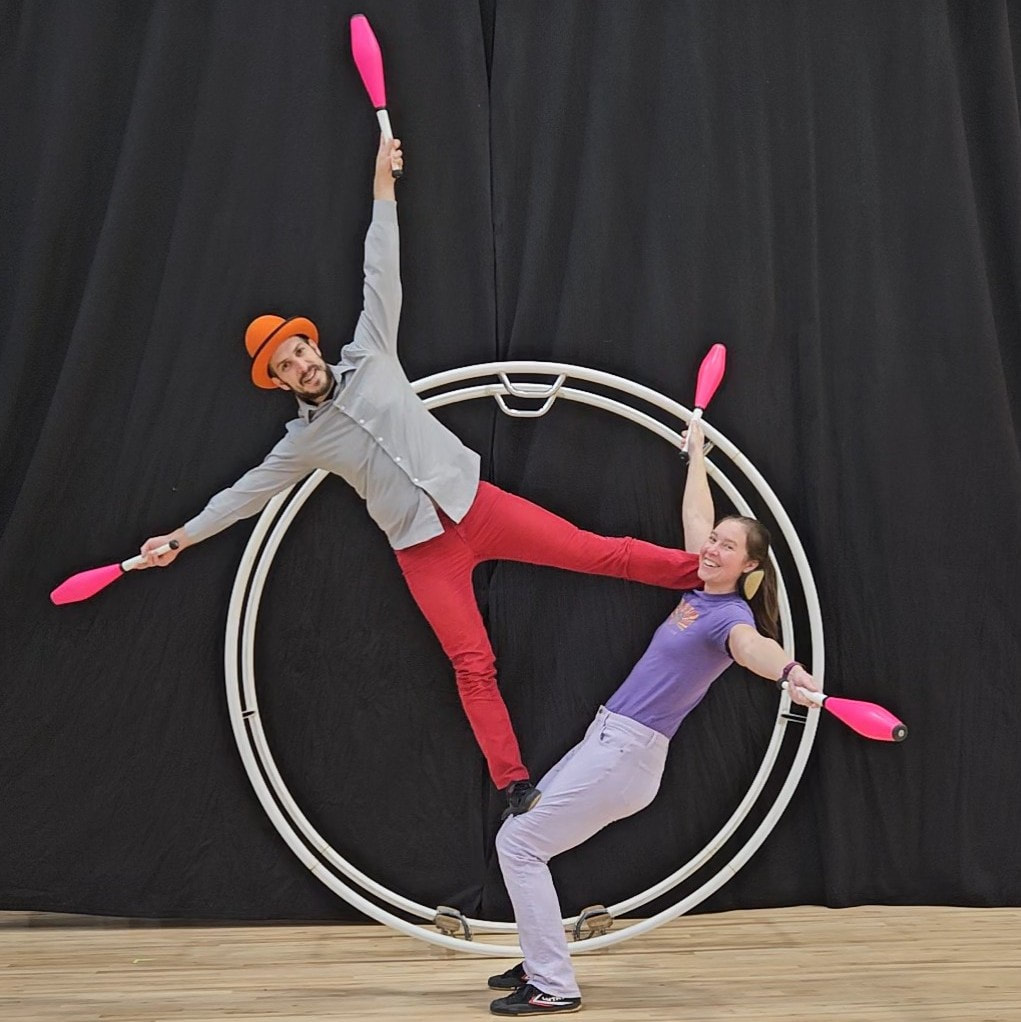 Two circus performers balance precariously inside a German wheel holding juggling pins.