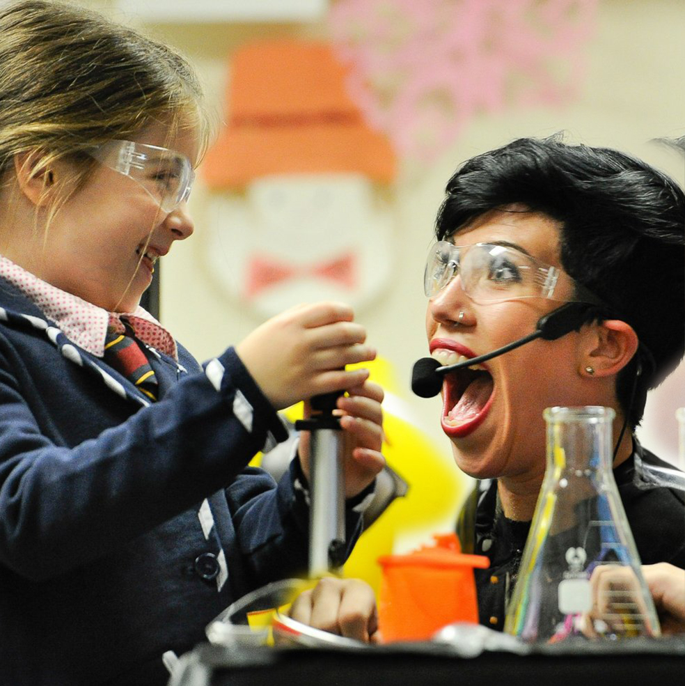 Young girl and a female science hero interact behind a table of beakers and other laboratory equipment. Huge smiles are on their faces.