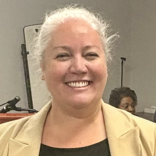 Woman with silver hair smiles. She's wearing a black shirt and tan blazer.