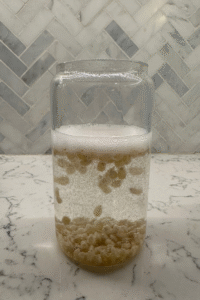 Clear jar with bubbling liquid and particles on a marble countertop with herringbone tile background.