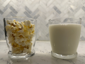 One glass filled with popcorn and one glass filled with milk sitting on a grey counter in front of a grey background